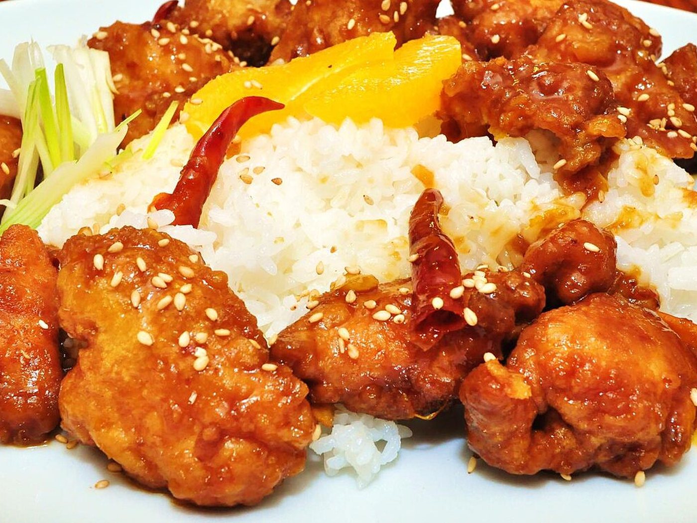 Glossy orange chicken topped with sesame seeds and sliced scallions in a white bowl